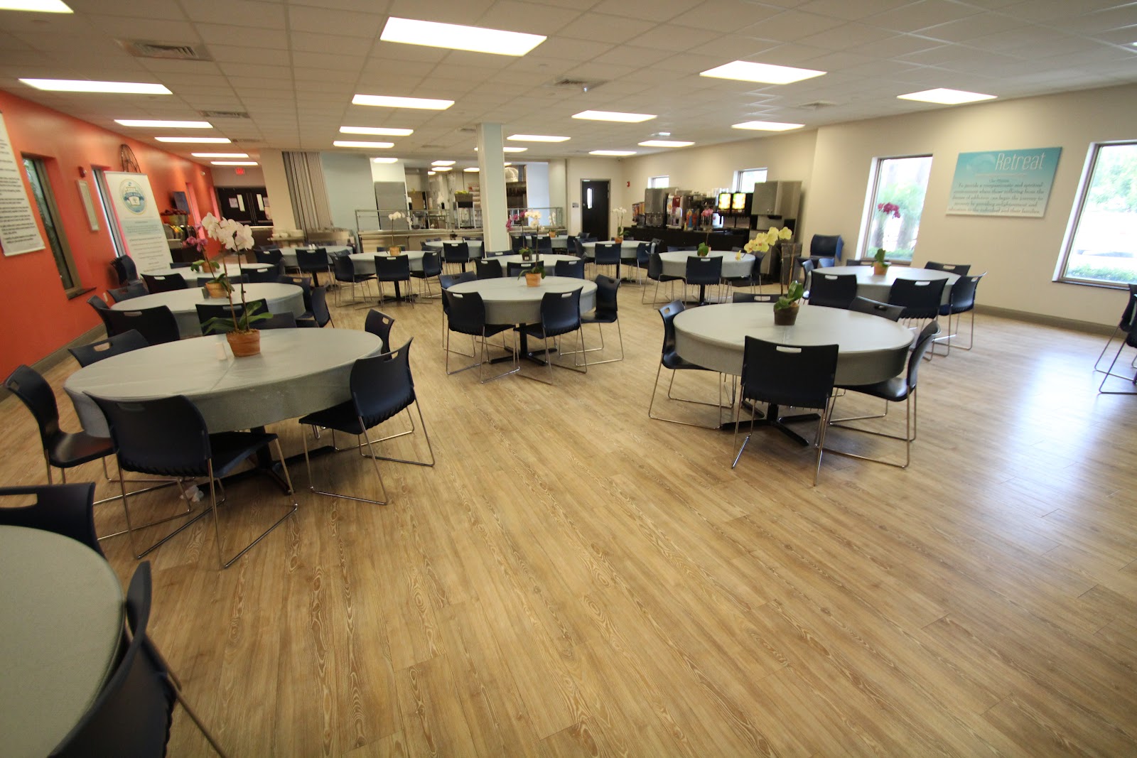 Interior large communal room with multiple round tables, black chairs, vibrant teal accent walls, artwork, and green plant displays