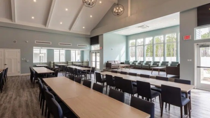 Interior dining and assembly space with long tables, chairs, high ceilings with skylights, and multiple seating areas