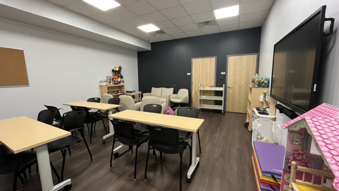 Interior activity room with tables, black chairs, shelving with games and supplies, TV screen, and seating areas