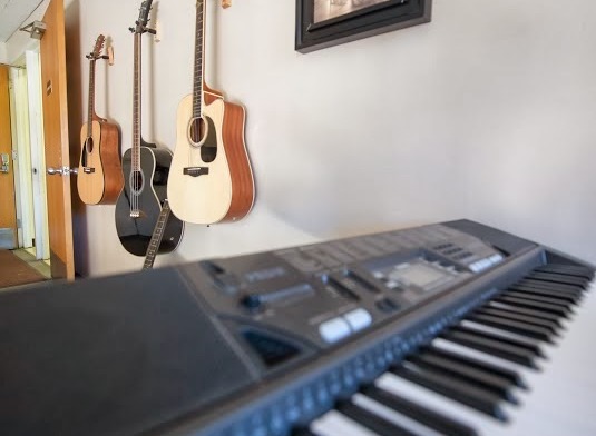 Interior room with keyboard instrument in foreground, multiple acoustic guitars on wall, and framed artwork