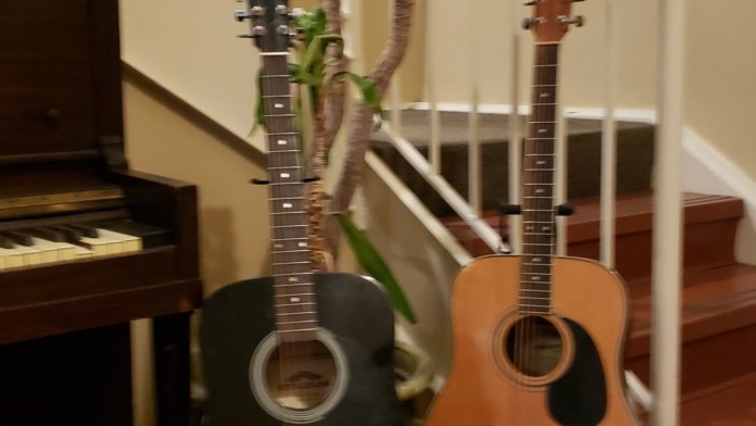 Interior recreation space with piano, two acoustic guitars on stands, spiral staircase with white railings, wall clock, and hardwood flooring
