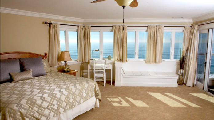 Interior spacious master bedroom with large bed, multiple windows with ocean vistas, seating area, crown molding, and coastal furnishings
