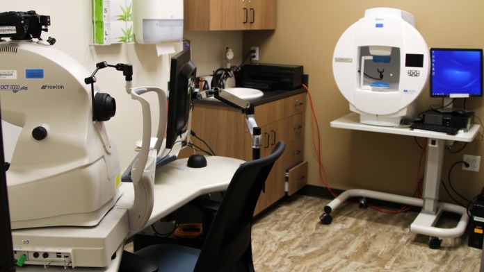 Interior eye examination room with specialized diagnostic equipment, patient chair, computer monitor, and medical instruments