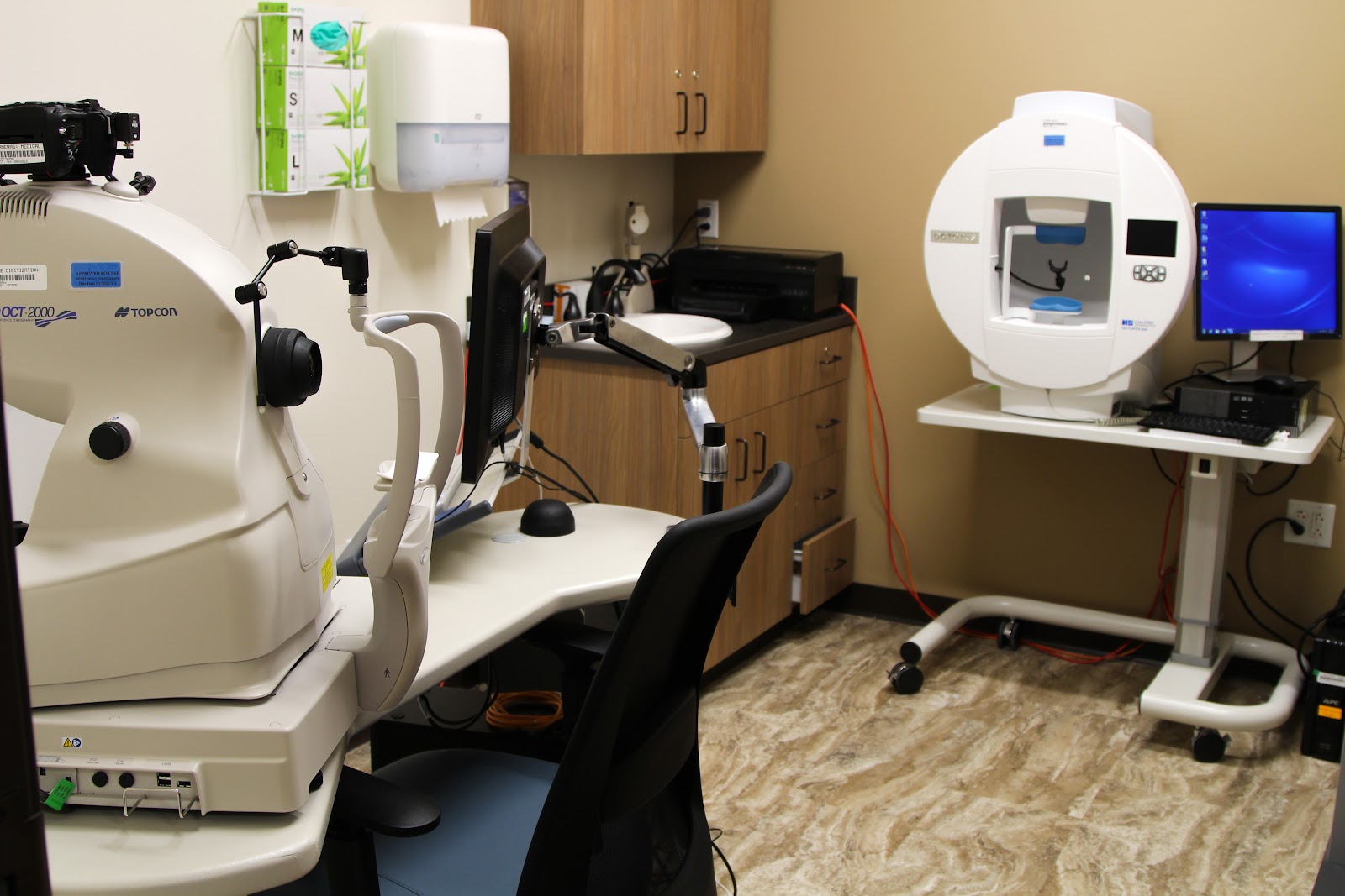 Interior eye examination room with specialized diagnostic equipment, patient chair, computer monitor, and medical instruments