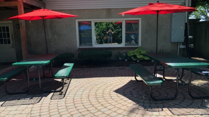 Patio space with red umbrellas, green picnic tables, brick pavement, and facility building backdrop