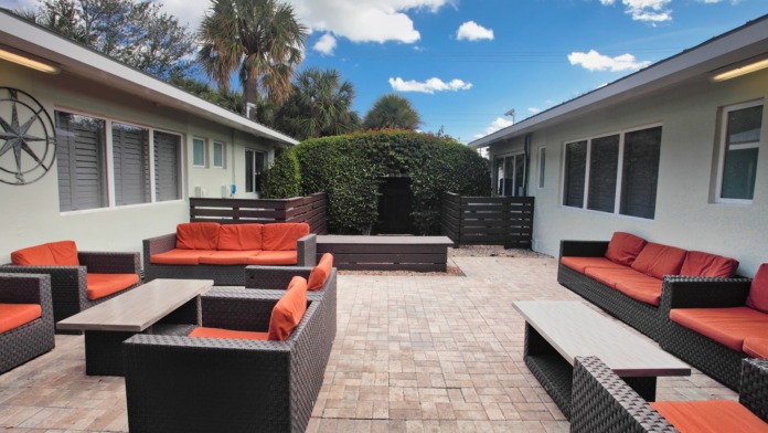 Outdoor courtyard space with woven black lounge furniture, orange cushions, coffee tables, and green hedge fencing.