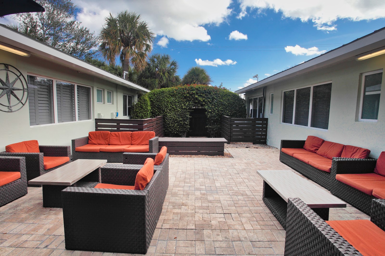 Outdoor courtyard space with woven black lounge furniture, orange cushions, coffee tables, and green hedge fencing.