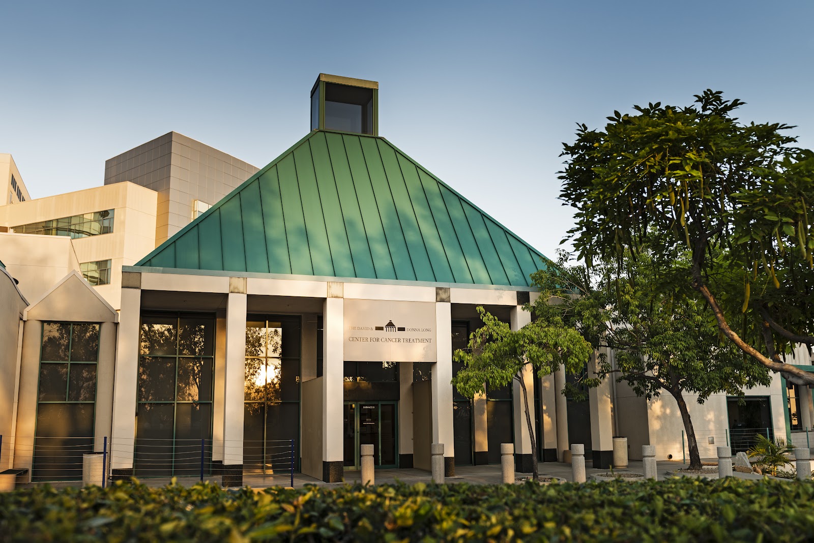 Exterior pavilion with green metal roof, white support columns, landscaping and trees creating facility entrance area