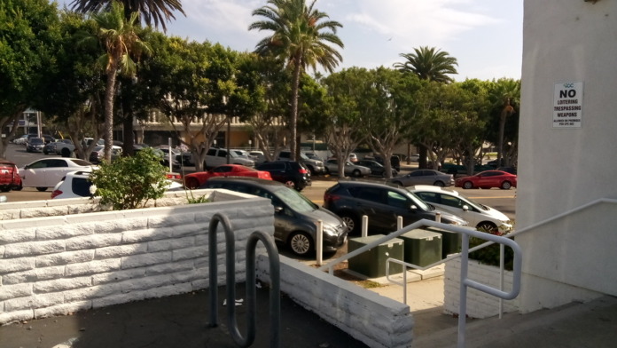 Exterior view showing parking lot with white building, palm trees, metal railing, and landscaping with accessible parking area