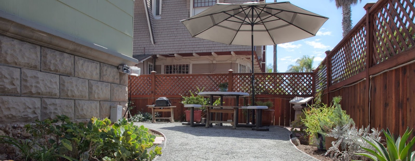Exterior patio space with dining table, large umbrella, stone walls, wooden lattice fence, and native plantings