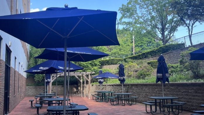 Exterior patio with multiple picnic tables, blue umbrellas for shade, brick flooring, building facade, and landscaped surroundings