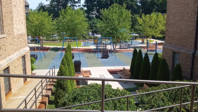 Outdoor view showing playground equipment, landscaped grounds with trees, brick building sections, and recreational areas