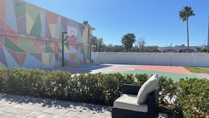 Exterior recreation area with colorful geometric mural, basketball court, seating, and landscaping under clear sky