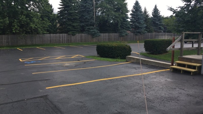 Exterior parking area with yellow parking space markings, green landscaping, wooden fence, and recreational structures in background