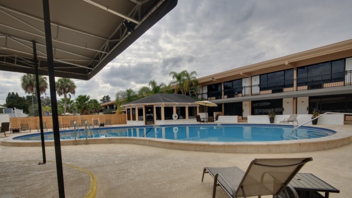 Exterior swimming pool with lounge chairs, palm trees, covered pavilion, and multi-story facility building in background