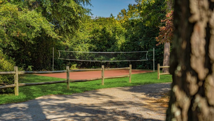 Exterior recreational area with volleyball net, sand court, wooden fencing, mature trees, and natural landscape