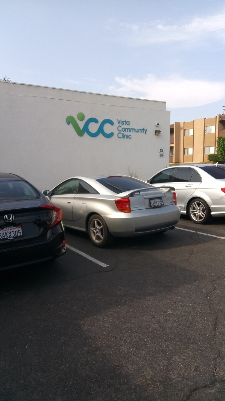 Exterior view of white building with Vista Community Clinic logo on wall and parked vehicles in lot
