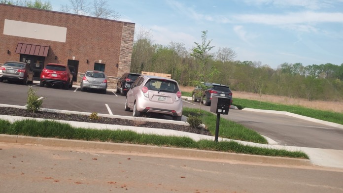 Exterior parking area with multiple vehicles and brick facility building with landscaping and grounds