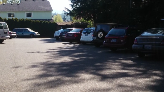 Exterior parking area with multiple vehicles and facility building visible with landscaping and privacy hedges
