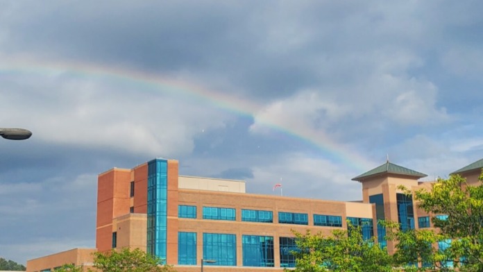 Exterior parking area with vehicles parked, turquoise windows of hospital building visible, landscaping, and cloudy sky