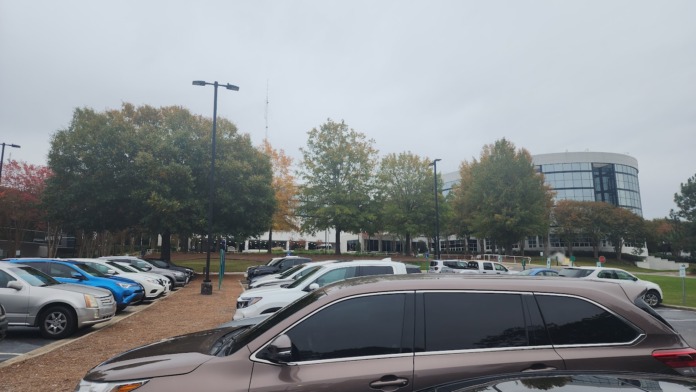 Exterior parking area with multiple parked vehicles, mature landscaping trees displaying fall colors, hospital building visible in background, clear sky