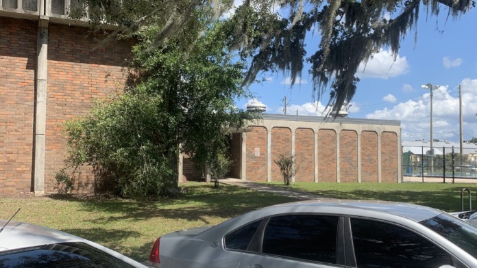 Exterior parking area with vehicles, brick building, mature oak tree with Spanish moss, and green lawn space