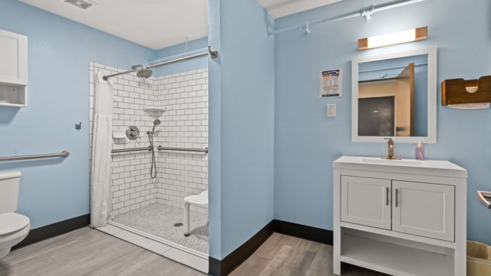 Interior bathroom with light blue walls, white subway tile shower with bench, white vanity cabinet with mirror, and gray flooring
