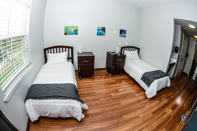 Interior residential bedroom with two twin beds, dark wood furniture, colorful wall art, natural window light, and hardwood flooring