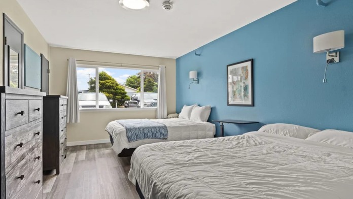 Interior patient bedroom with two beds, blue accent wall, dresser with mirror, windows, and wood-look flooring