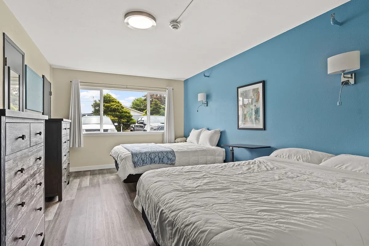 Interior patient bedroom with two beds, blue accent wall, dresser with mirror, windows, and wood-look flooring