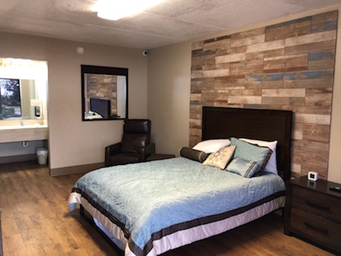Interior patient bedroom featuring queen bed with blue striped bedding, wooden accent wall, nightstand, desk, and ensuite bathroom