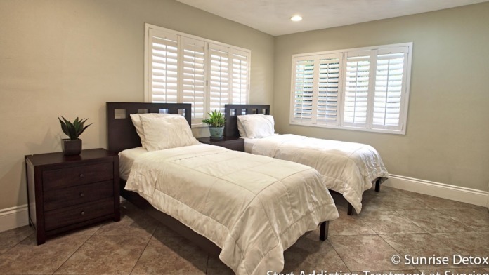 Interior patient bedroom with two twin beds, white bedding, dark wood headboards, nightstands, and shuttered windows.