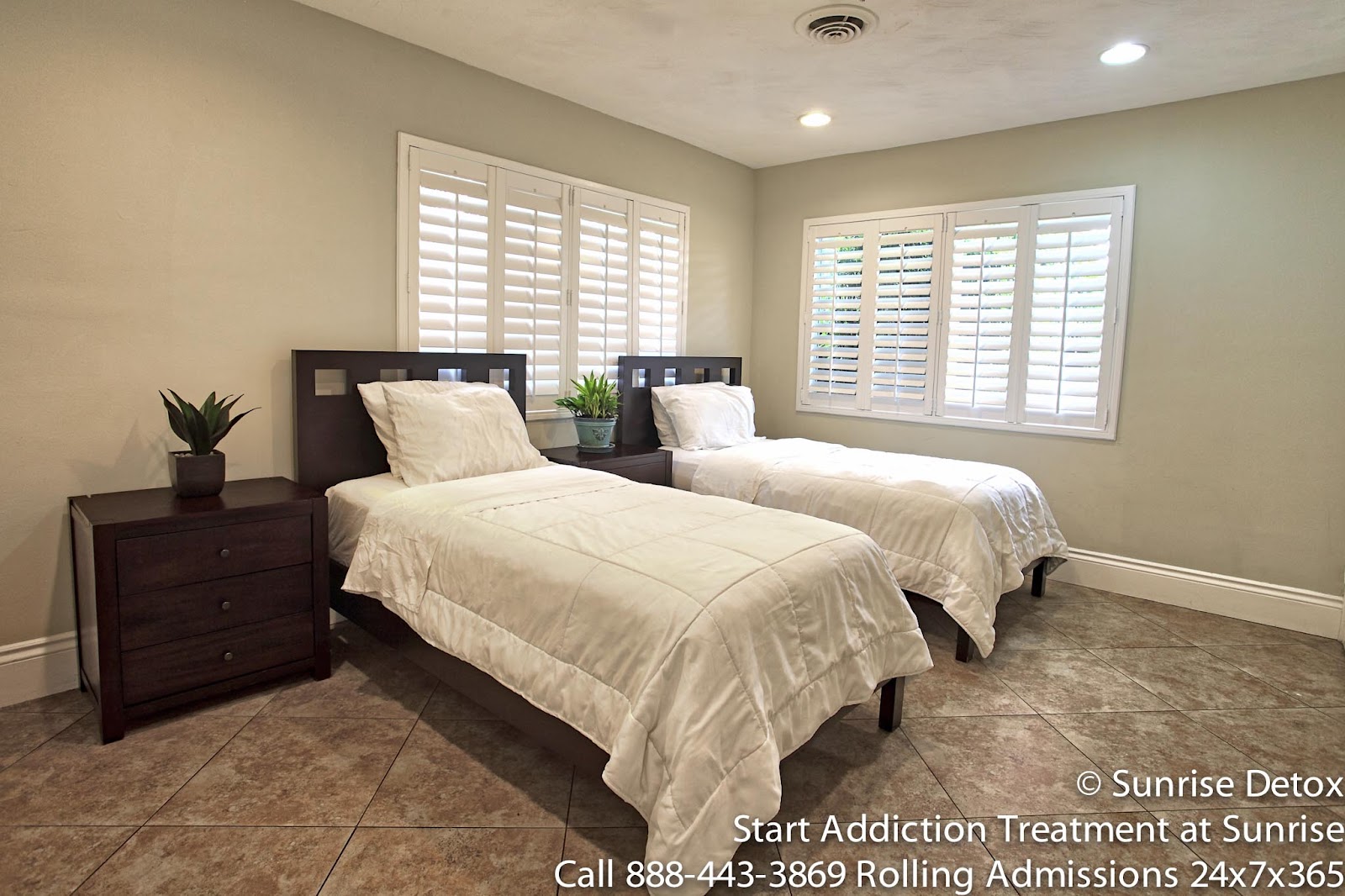 Interior patient bedroom with two twin beds, white bedding, dark wood headboards, nightstands, and shuttered windows.