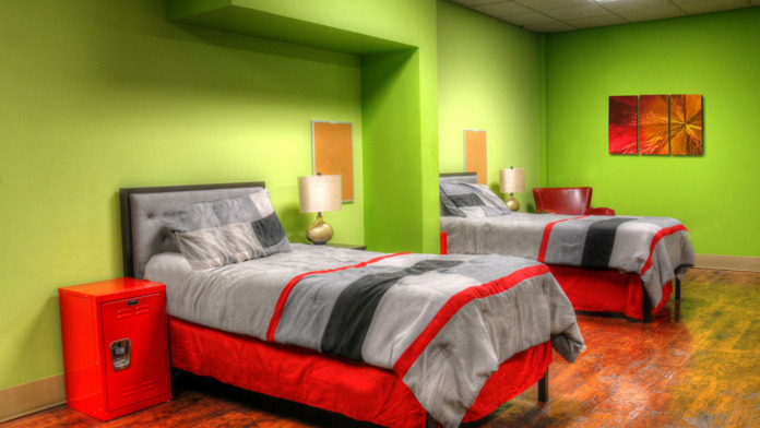 Interior bedroom showing twin beds with red and striped bedding, nightstand with lamp, red storage locker, and lime green accent walls with artwork