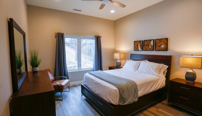 Interior bedroom featuring queen bed with white linens, nightstands with lamps, desk, vanity mirror, and large window