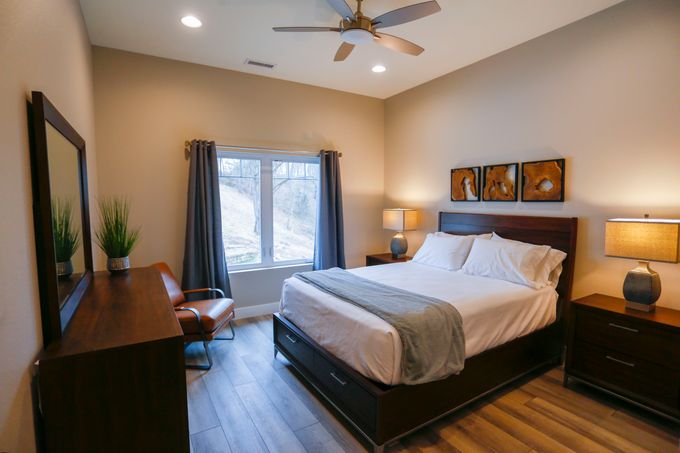 Interior bedroom featuring queen bed with white linens, nightstands with lamps, desk, vanity mirror, and large window
