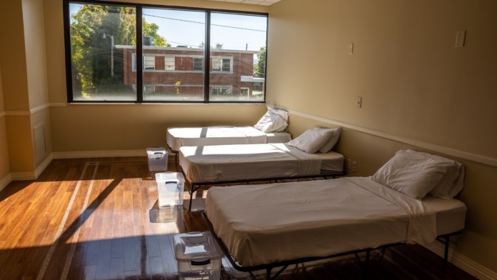 Interior patient bedroom featuring three single beds with white linens, hardwood flooring, and large windows with natural light