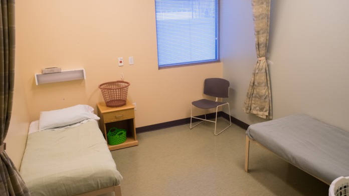 Interior patient bedroom featuring two twin beds with linens, nightstands, laundry basket, chair, window with blind, and closet space