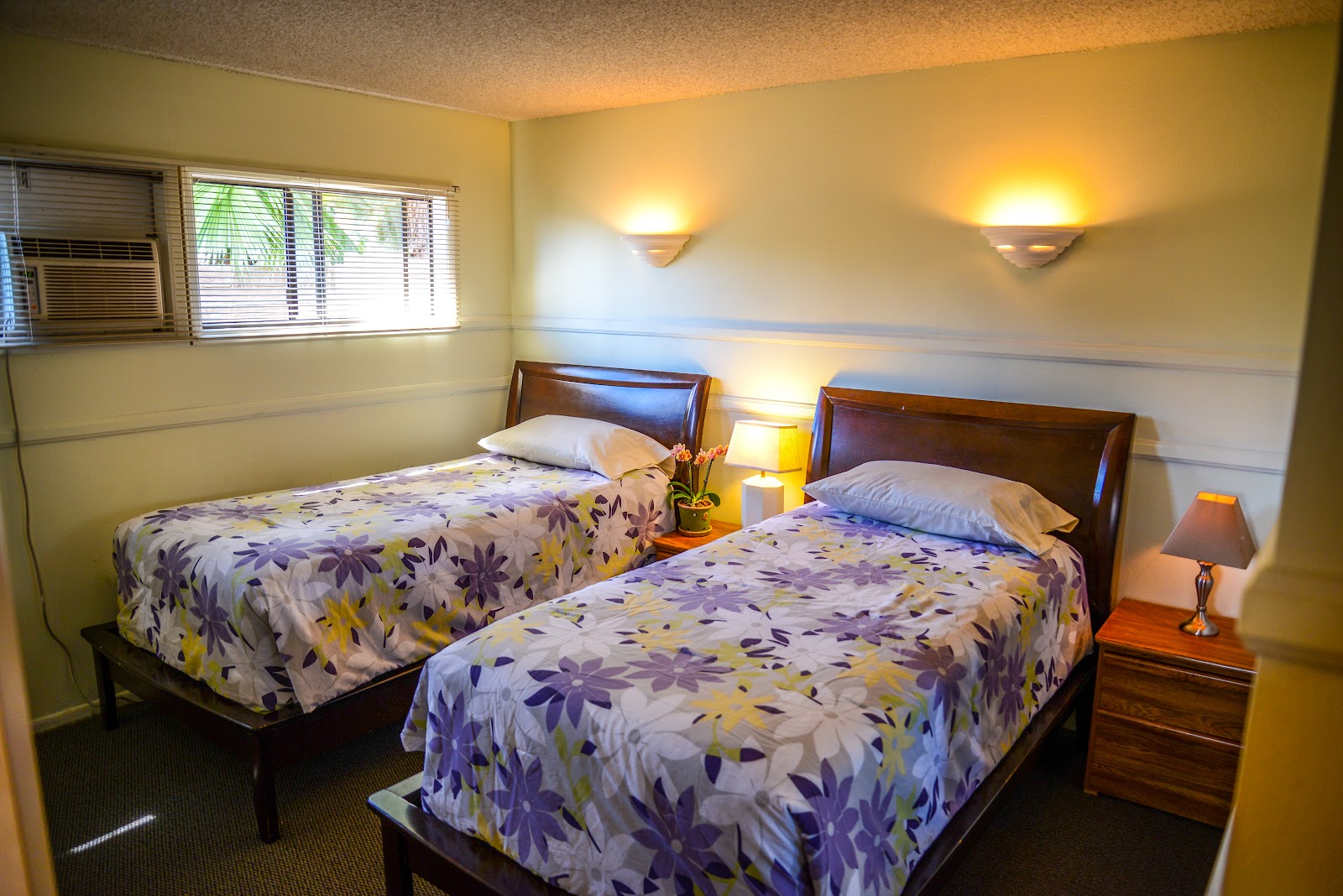 Interior patient bedroom featuring two twin beds with floral comforters, wood furniture, nightstands with lamps, and window with blinds