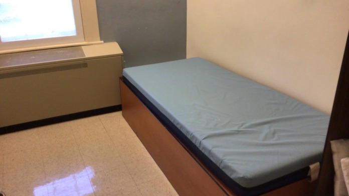 Interior patient bedroom showing single bed with mattress, window with natural light, and minimal furnishings