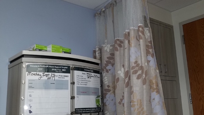 Interior patient room corner with wall-mounted information boards, tissue dispenser, and patterned fabric privacy curtain suspended from ceiling track system