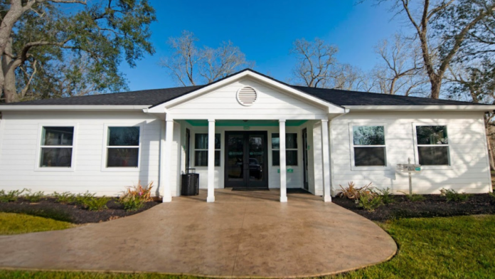 Exterior of single-story white cottage building with green accents, covered entry porch, windows, and landscaped grounds