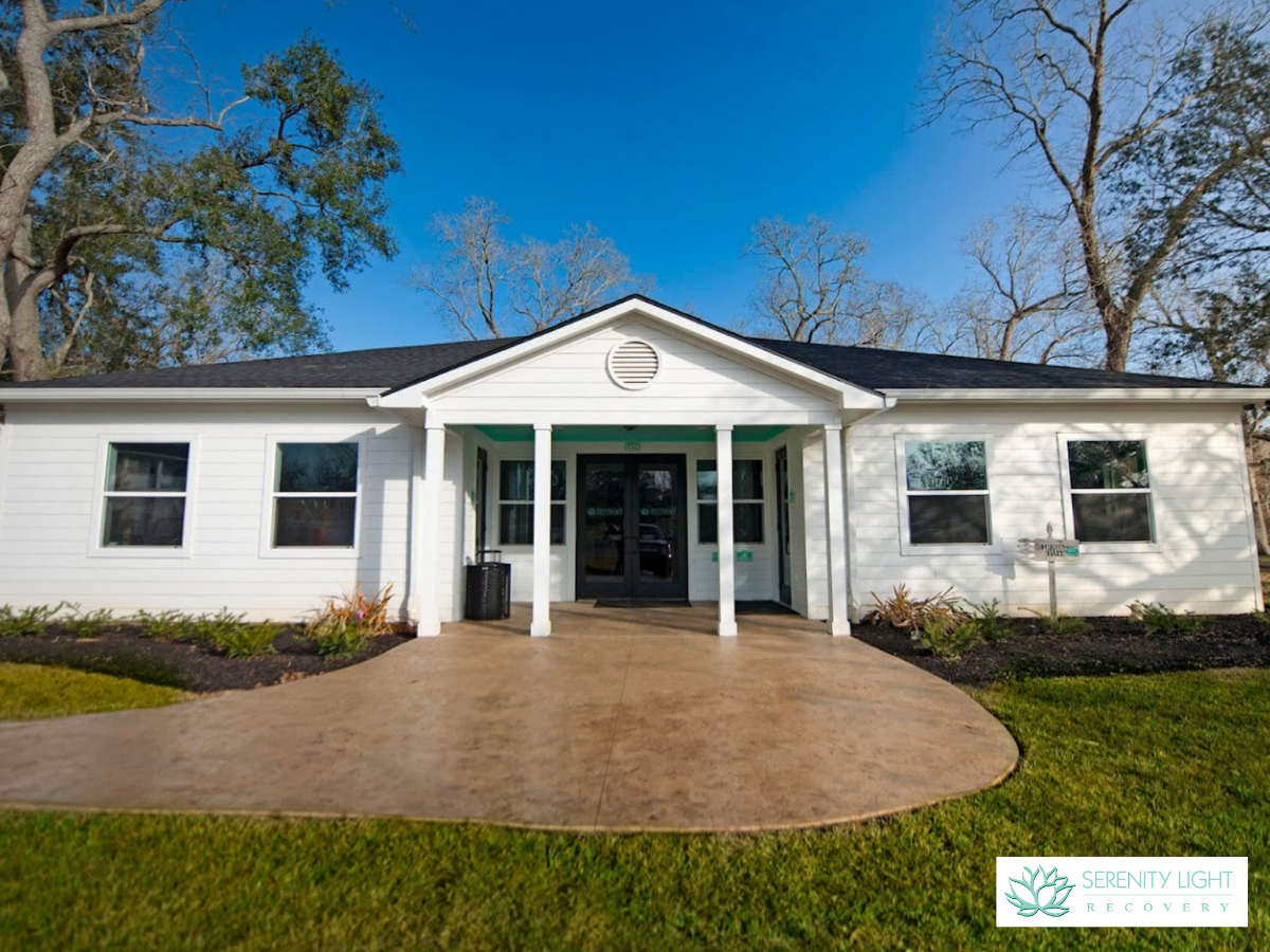 Exterior of single-story white cottage building with green accents, covered entry porch, windows, and landscaped grounds