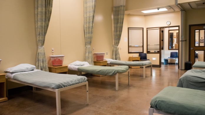 Interior patient room with multiple single beds, bedside tables, laundry baskets, windows with curtains, and neutral walls