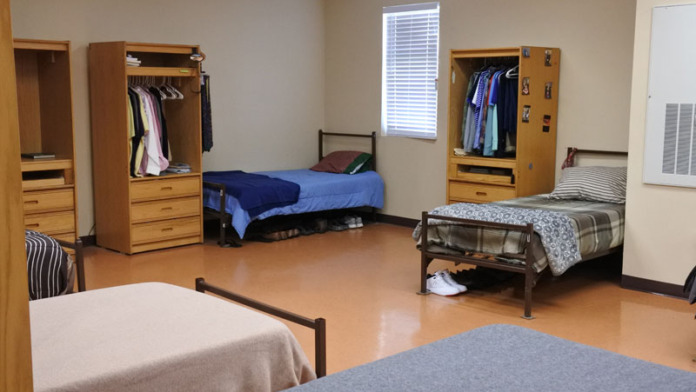 Interior dormitory room with multiple beds, wooden wardrobes, windows, and personal storage areas