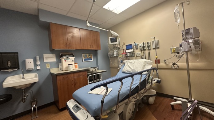Interior medical examination room with blue exam table, wood cabinetry, wall-mounted monitors, sink, and medical equipment storage