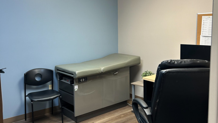 Interior examination room with medical table, black chair, cork board with documents, cream walls, and wooden flooring.
