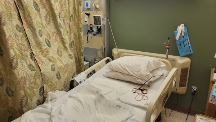 Interior patient bedroom showing hospital bed with white bedding, medical monitoring equipment, oxygen outlet, and patterned privacy curtain