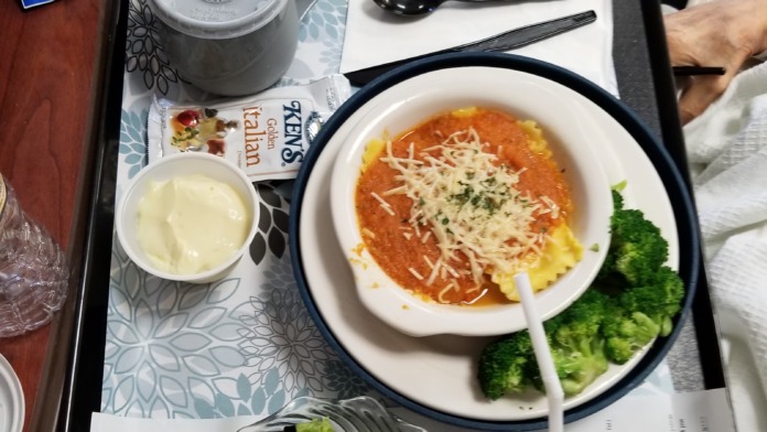 Interior close-up of patient meal tray with prepared dish, salad, beverage, and disposable serviceware on bedside table