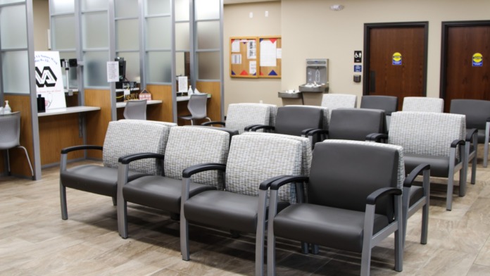 Interior waiting room with multiple upholstered chairs, reception desk with glass partitions, and water station